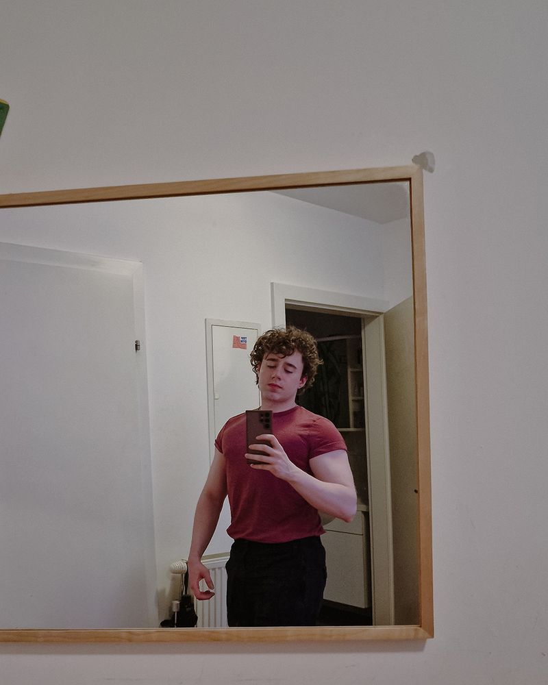 Selfie of a guy, curly hair, red shirt, slightly flexing his arms and chest