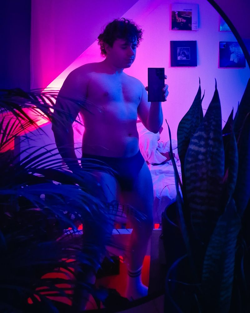 Muscular guy in undies posing in front of the mirror in gloomy blue-pink lighting.