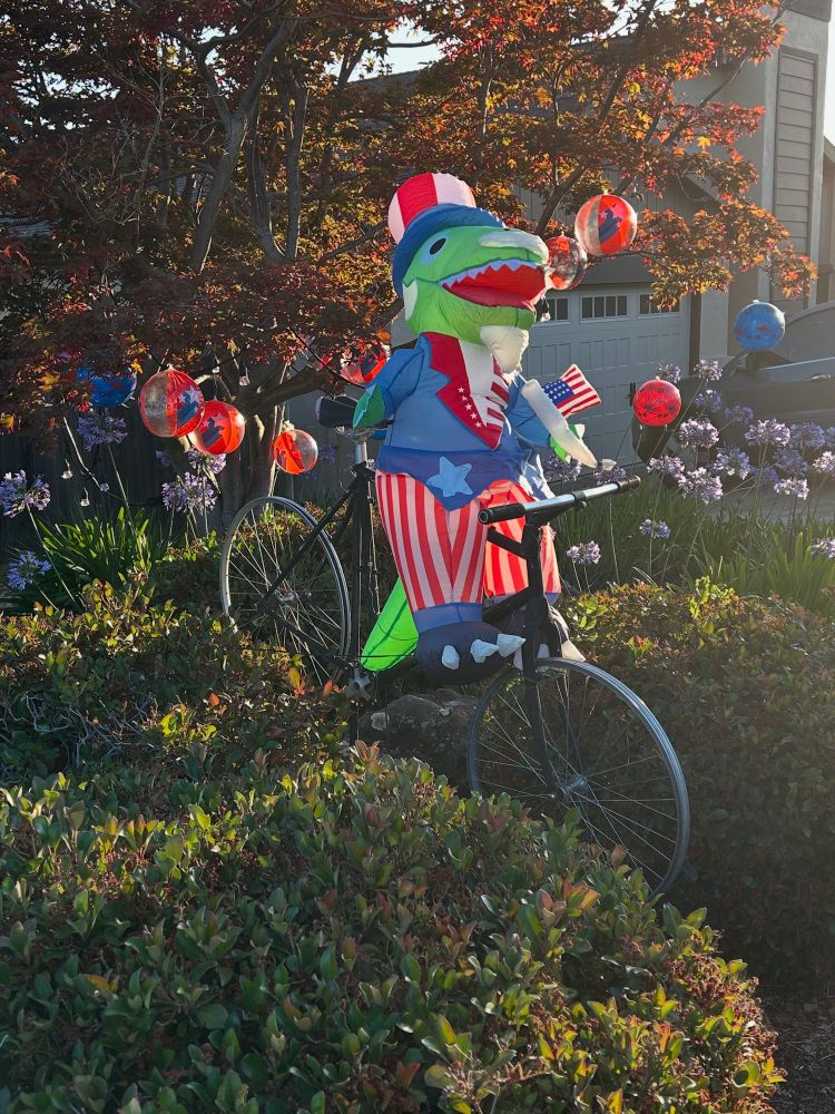 A T. rex dressed as Uncle Sam holding an American flag and riding a bicycle.