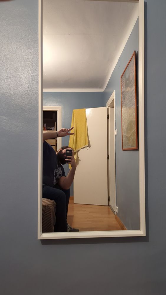 foto de un espejo en el que se me ve reflejado sentado en la cama, delante de mí hay un cuadro y al fondo una puerta abierta con una toalla colgando 