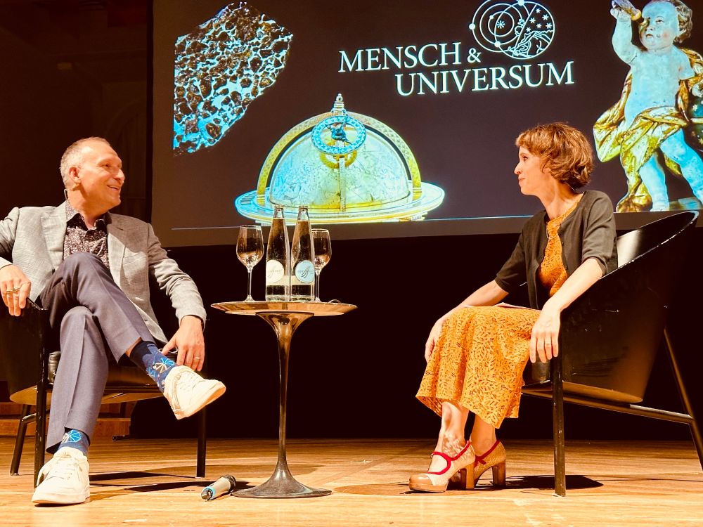 Astrophysicist Thomas Zurbuchen and philosopher Barbara Bleisch sitting together and discussing, laughing, on a podium in St. Gallen, Switzerland. In the background, a title image says "Mensch & Universum" (Humanity & Universe).