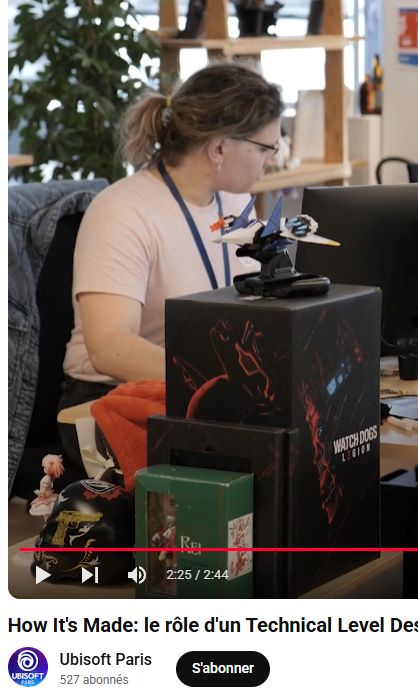 me from two years ago, not blonde yet, hair in a poneytail, pink shirt, working at my desk, half hidden behind many goodies/merch