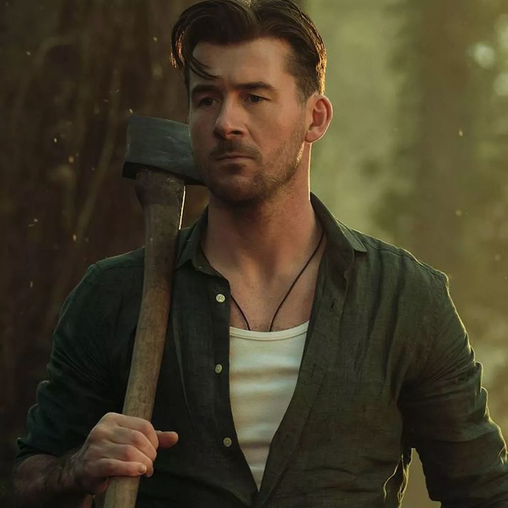 Barry Sloane wearing a tank top under a loose linen shirt with many buttons open and an axe slung over his shoulder smouldering. 