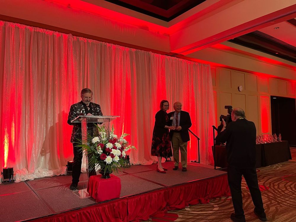 Stage with one person standing at a podium, two people standing at the right, photographer in foreground