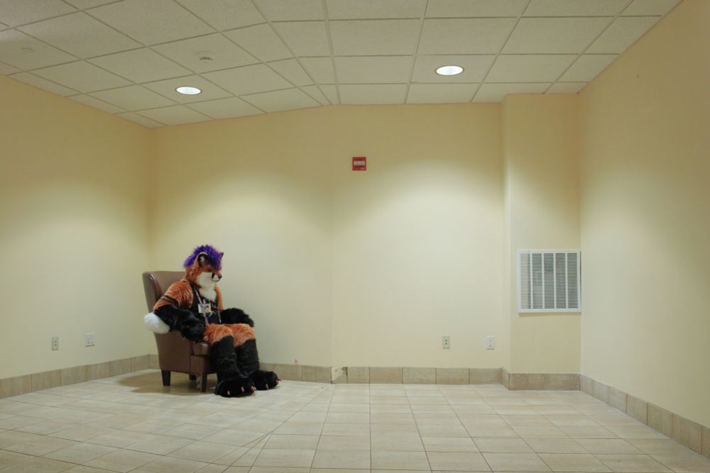 A fox fursuiter sitting in a chair in an empty hall. The wall has an angle to it and the tile in the floor does not match up perfectly as a result. There's a broken tile right at the angle in the wall, a grate to the far right, a fire strobe high in the middle, and several wall plates.