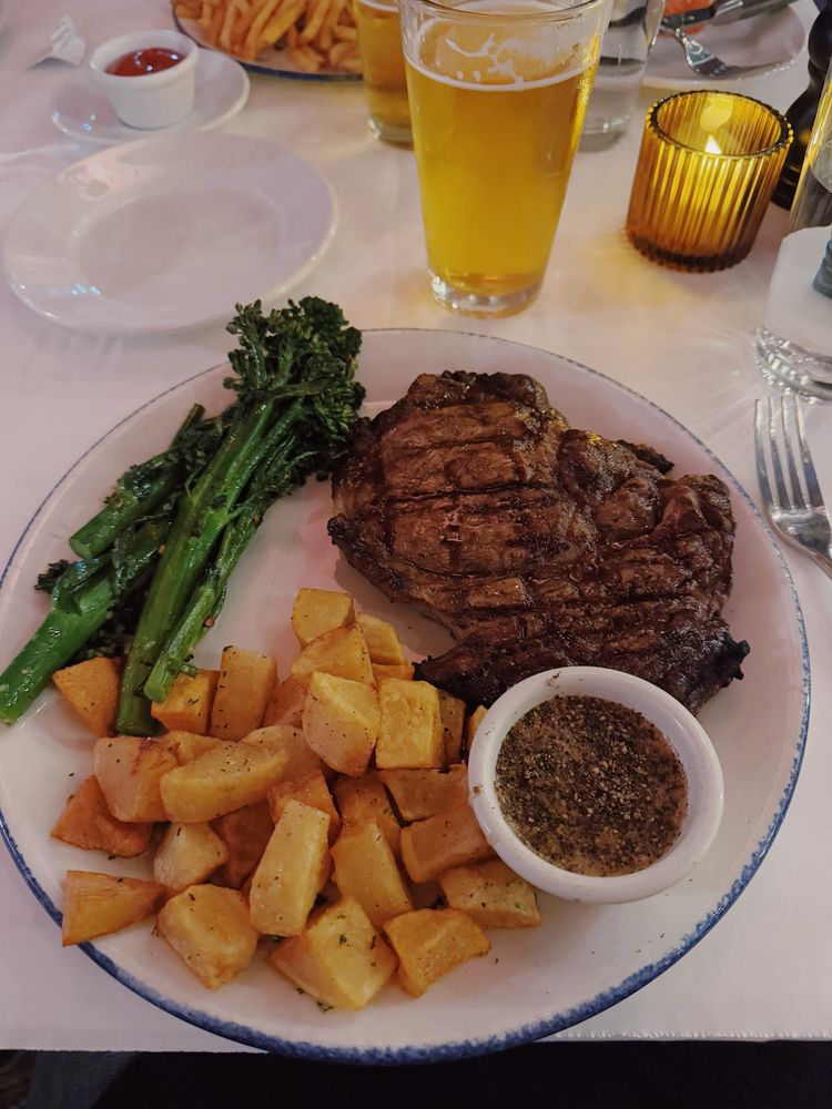 Plated rib eye with broccolini, papas bravas, and pepper sauce. There is a beer and candle on the table. 