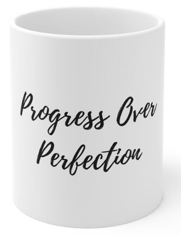 Plain white mug with black writing that reads "Progress Over Perfection"