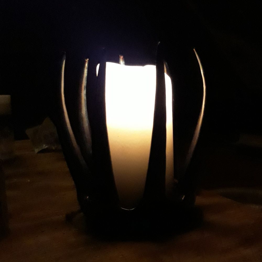 A very dark picture of a horned black candle holder, looking somewhat like a crown, with a lit thick white candle in the middle