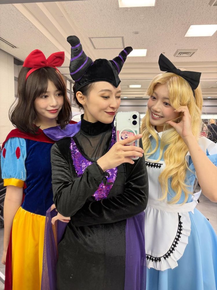 A picture of Yuzuha Saeki, Miu Suzuki, and Mirai Hinata dressed as Snow White from Snow White and the Seven Dwarfs, Maleficent from Sleeping Beauty, and Alice from Alice in Wonderland. Miu is taking the picture with her phone as they all smile. Mirai is making a heart expression with her left hand.