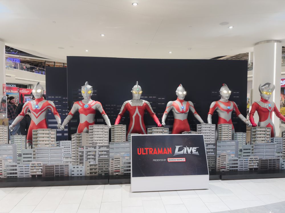 A picture of a bunch of Ultraman suits standing next to each other in a row. From left to right is Zoffy, Ultraman Ace, Ultraseven, Ultraman, Ultraman Jack, and Ultraman Taro. In front of them are buildings, with a sign that says "Ultraman Live, presented by Bandai Namco".