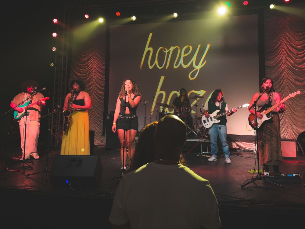 A picture of the band Honey Haze playing on stage at Big and Tall Fest. From left to right, Ethan Govea on guitar, Alondra on saxophone, Alyssa on lead vocals, Myles Yambao on drums, Jenni on bass, and Ella on guitar and backing vocals. Behind them is the Honey Haze logo.