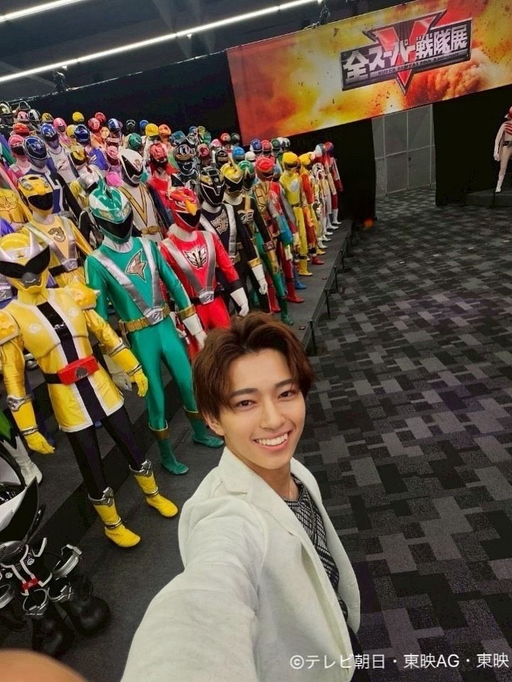 A picture of the actor Hideharu Suzuki taking a selfie near a bunch of mannequins wearing various ranger suits throughout the Super Sentai series. Hideharu is dressed in the clothing of the character he plays in No.1 Sentai Gozyuger, Rikuo Byakuya. There's a banner in the back that features the Super Sentai 50th Anniversary logo with an explosion in the background. The text reads "全スーパー戦隊", which translates to "All Super Sentai". Underneath the Japanese text reads "Super Sentai 50th Anniversary". Behind the all the text is a giant "V", which is the Super Sentai logo, but instead of the V being in gold, it's in red. There's text on the bottom right of the photo that says "©テレビ朝日·東映AG·東映", which translates to "©TV Asahi · Toei AG · Toei".