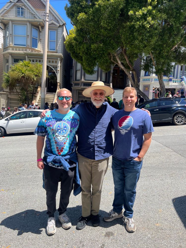 Three men posing in front of a San Francisco Victorian. The one in the center is noticeably older, wearing a sun hat and with glasses and a beard. The two younger men are both wearing Grateful Dead t-shirts:
