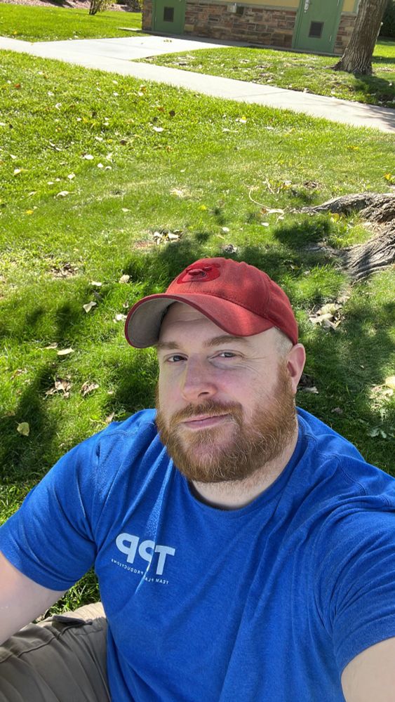 Account owner selfie while sitting on grass in shade of a tree. Blue shirt, red hat.