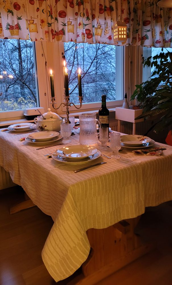 Katettu pöytä kynttilöineen odottaa ruokavieraita. Ikkunan takana hämärtyy ilta. 