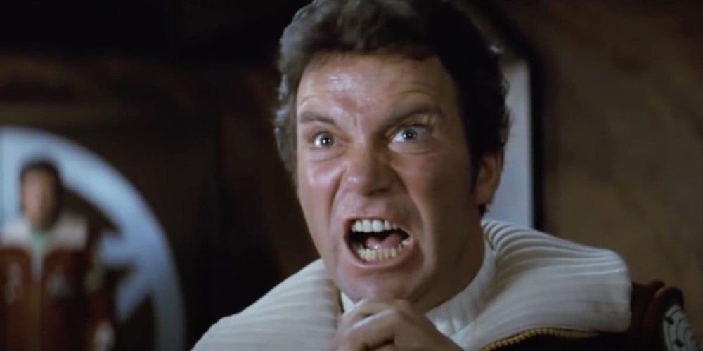 A man (Star Trek TOS: James T. Kirk) in a white and red uniform is shown mid-shout with an intense, angry expression, eyes wide and teeth bared. His hands are clenched near his face. The scene is indoors with a blurred figure in the background. The image is a famous moment from the film Star Trek II: The Wrath of Khan.