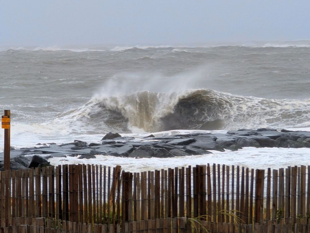 A large wave crashes dramatically over a rock jetty, with wind-driven spray rising into the air. The ocean appears rough and turbulent, with layers of choppy waves extending to the horizon under a gray, misty sky. In the foreground, wooden sand fencing and a yellow warning sign line the edge of the beach, emphasizing the hazardous coastal conditions caused by Hurricane Erin.