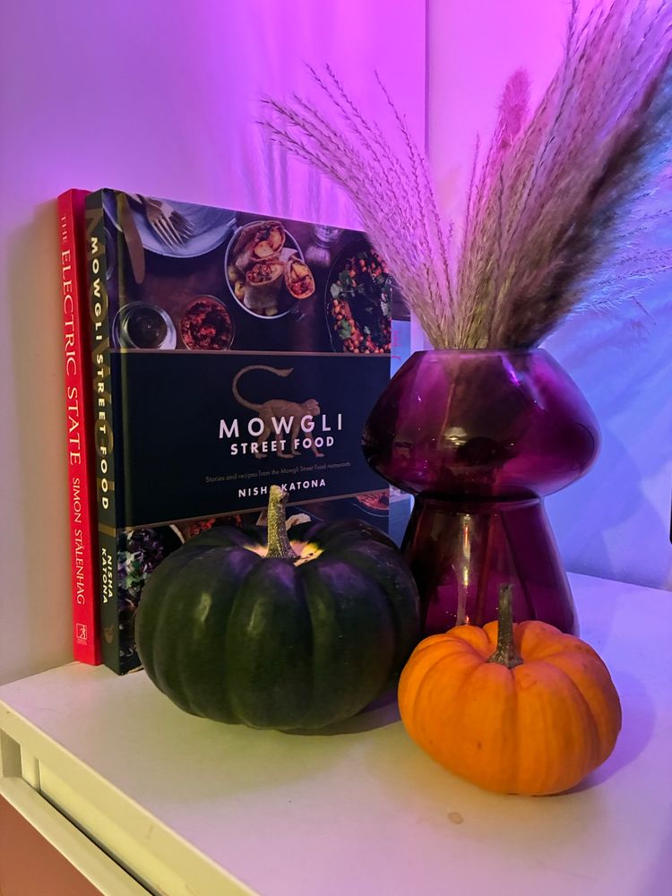 A picture of two small pumpkins, the slight bigger one is a very dark, almost black green and the smaller one is orange. Behind them is a mushroom shaped purple glass vase with pampas grass. Next to them are two books, a mowgli street food cook book and the electric state. 