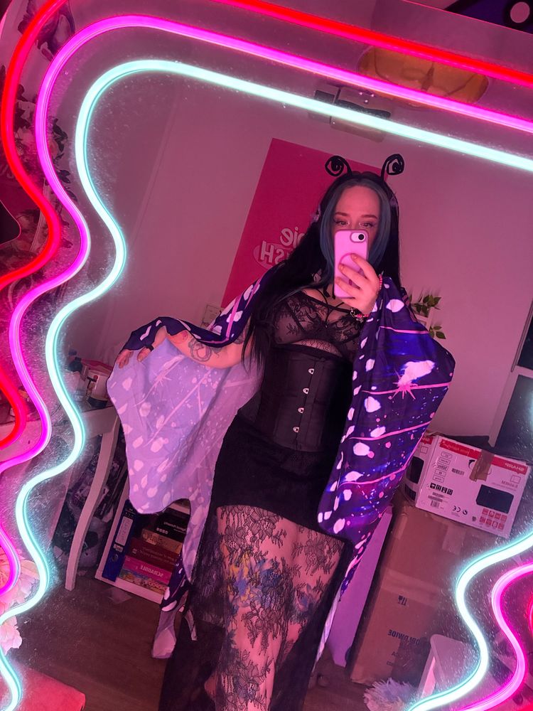 A mirror selfie of my wearing a netted black dress with black bodycon skirt and black corset. I have on purple and blue long fabric butterfly wings and a headband that looks like curly little antenna. 