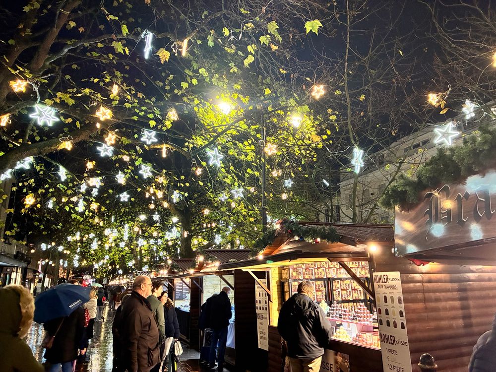 A picture of a Christmas market. It’s night time, and the trees are filled with yellow and white star shaped fairy lights. There are wooden cabins filled with miscellaneous trinkets, the mist visible one has assorted flavoured chocolates. There’s people milling around and viewing goods at the stalls. 