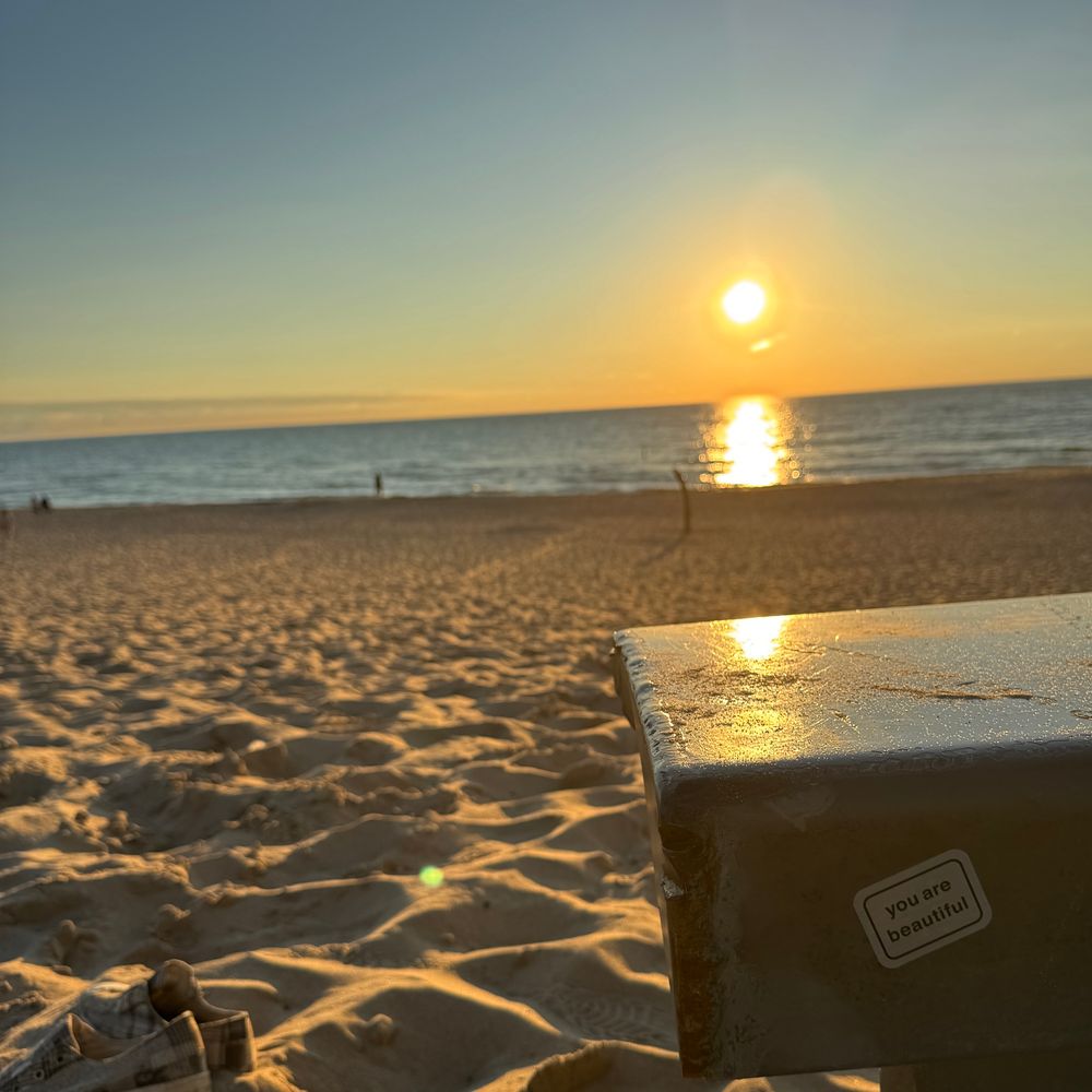 sun setting over water and a sandy beach with a sticker that says "you are beautiful" on a metal support in the foreground

