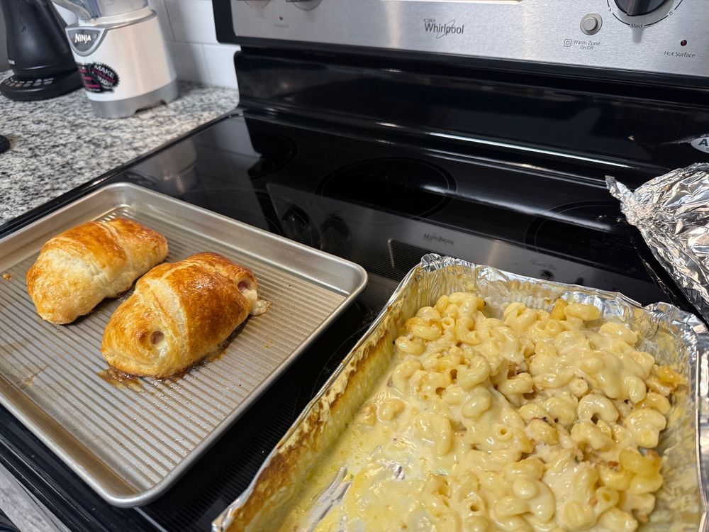Stove top with 2 pans. The first has 2 ham and cheese croissant sandwiches with an egg wash. The second has leftover Mac n cheese casserole 