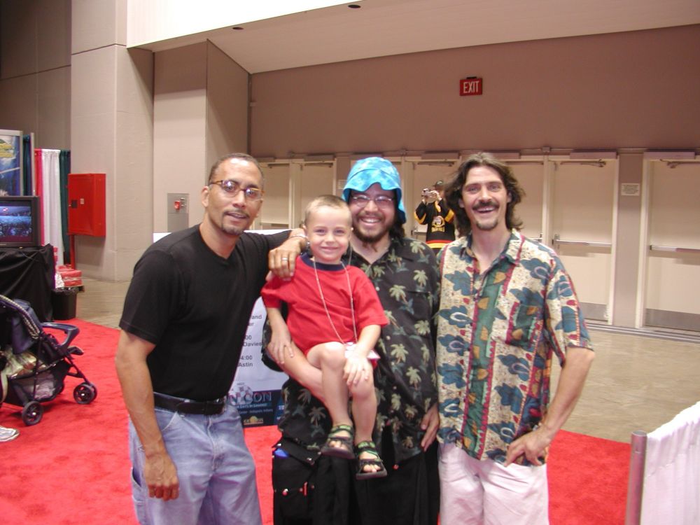 The actors Richard Biggs and Jason Carter standing next to a man in a black kilt, black aloha shirt with palm trees and a floppy blue tie dye hat. He is bearded, wearing glasses and holding a small child in his arms. The child has close shaven hair, a red shirt, strappy sandles and some sort of necklace. They are all standing in a convention center.