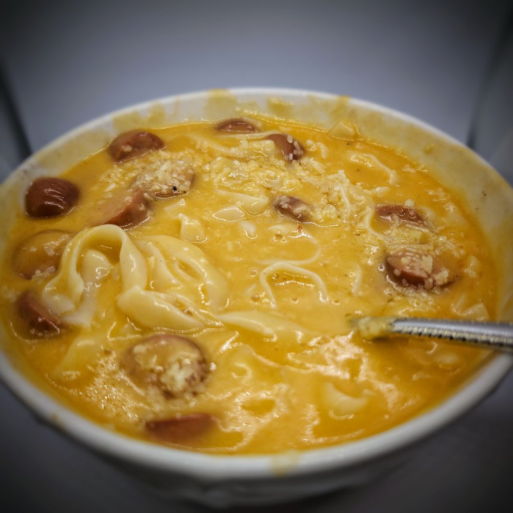 A white bowl filled with and orange cheese sauce, long noodles and small, circles of hot dog.