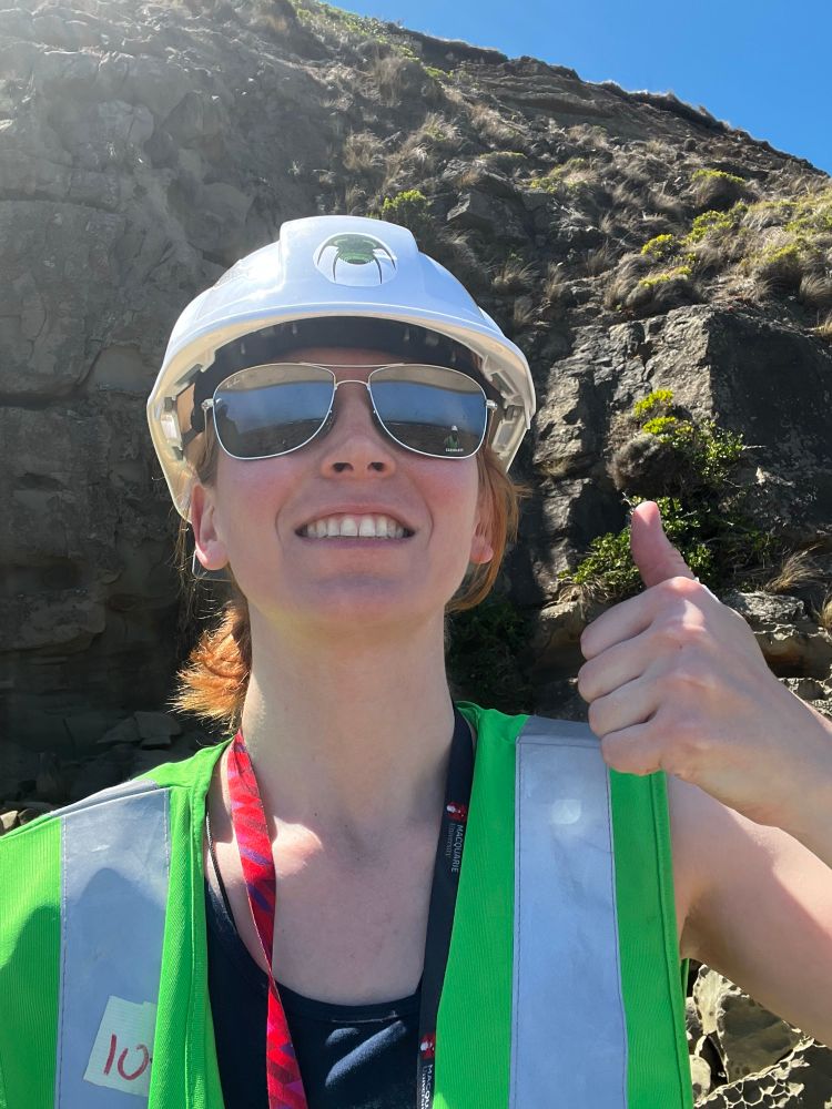 A strangely proportioned smiling lady (why is her neck so long??) with a hi vis vest and a hard hat gives a thumbs up. She’s outside, where it’s sunny and nice, and also there are lots of rocks!