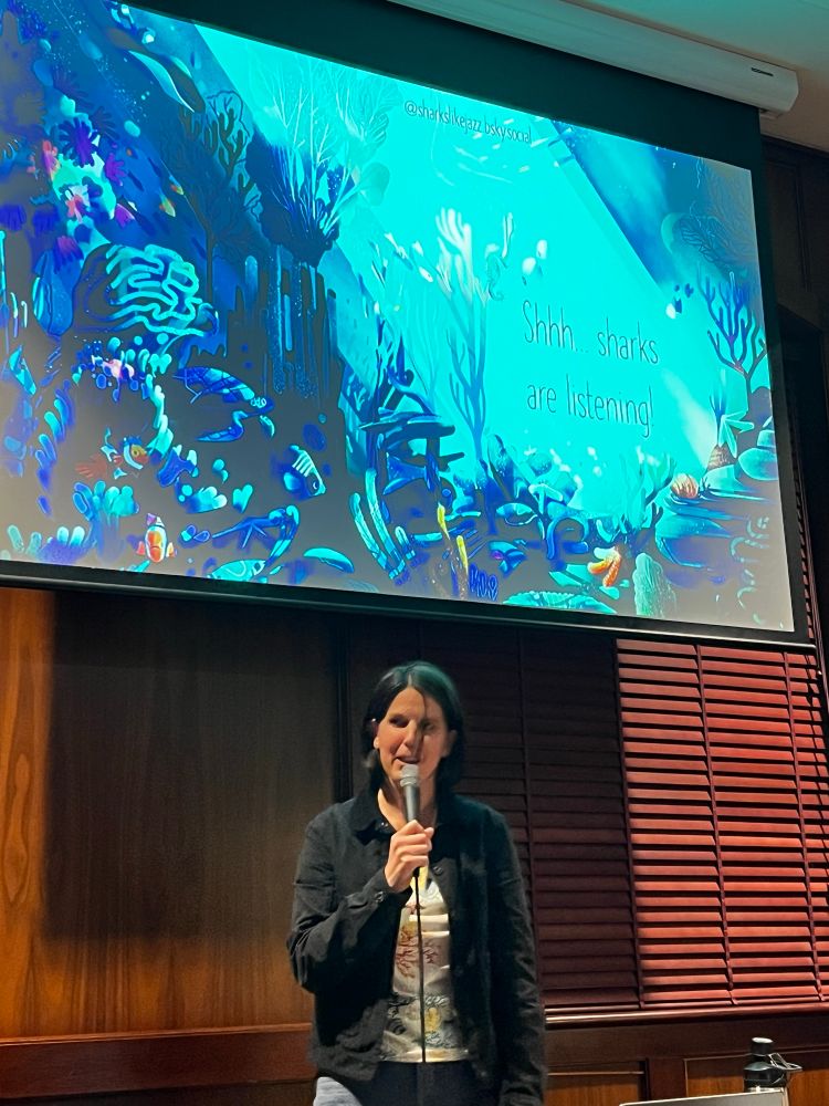 A confident woman stands before an ocean themed presentation, speaking calmly into the microphone. It’s Lucille!