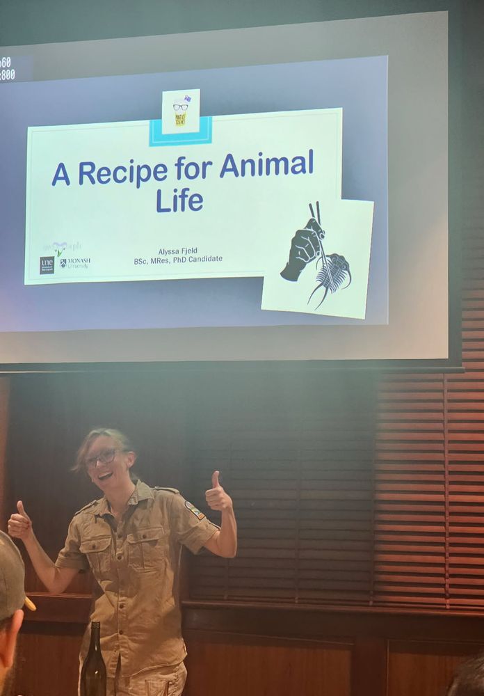 A woman in some kind of paleontologist get up gives a double thumbs up, standing in front of a powerpoint screen. Her smile is large and unsettling. It’s me!