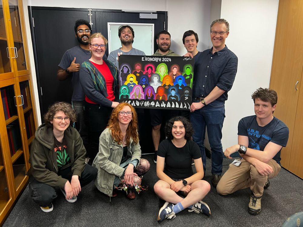 A smiling lab group poses with a large banner, depicting vectorized silhouettes of 17 evolutionary morphology researchers.  Astrid, the artist, is holding the left hand side of the banner!