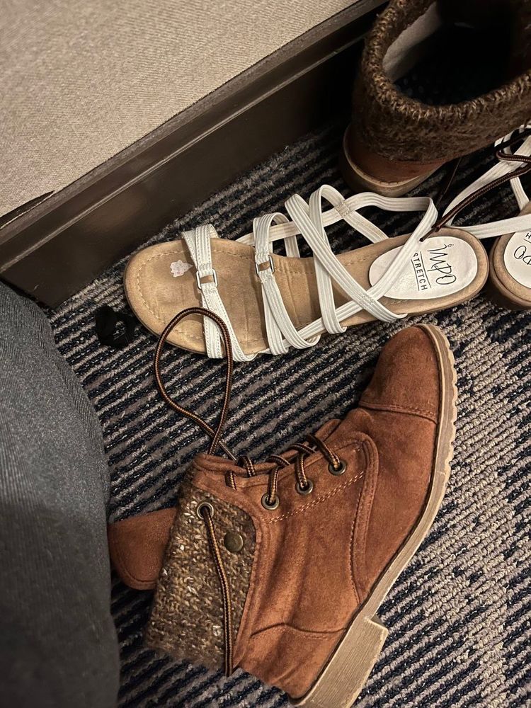 Brown boots and some sandals with white straps lay messily on the floor. In the toe area of one of the sandals there is a worn down sticker. The design is not recognizable.