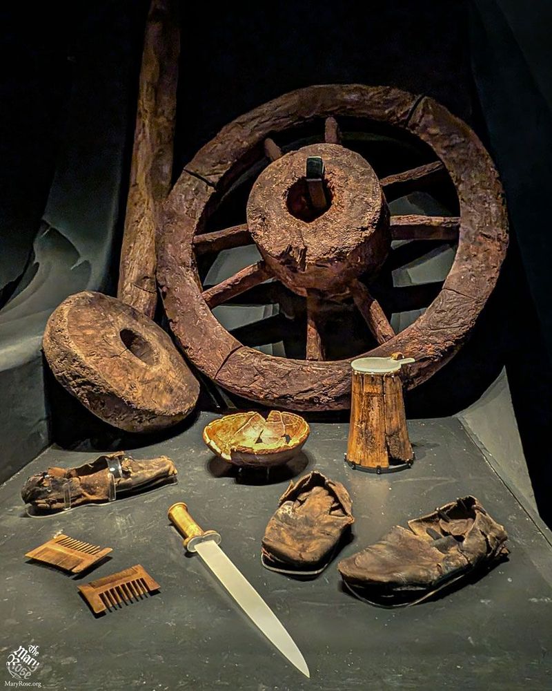 A spoked wooden gun carriage wheel propped up against a wall, surrounded by items including a dagger, nit combs, shoes and a flagon. There is also a smaller solid wheel next to it.