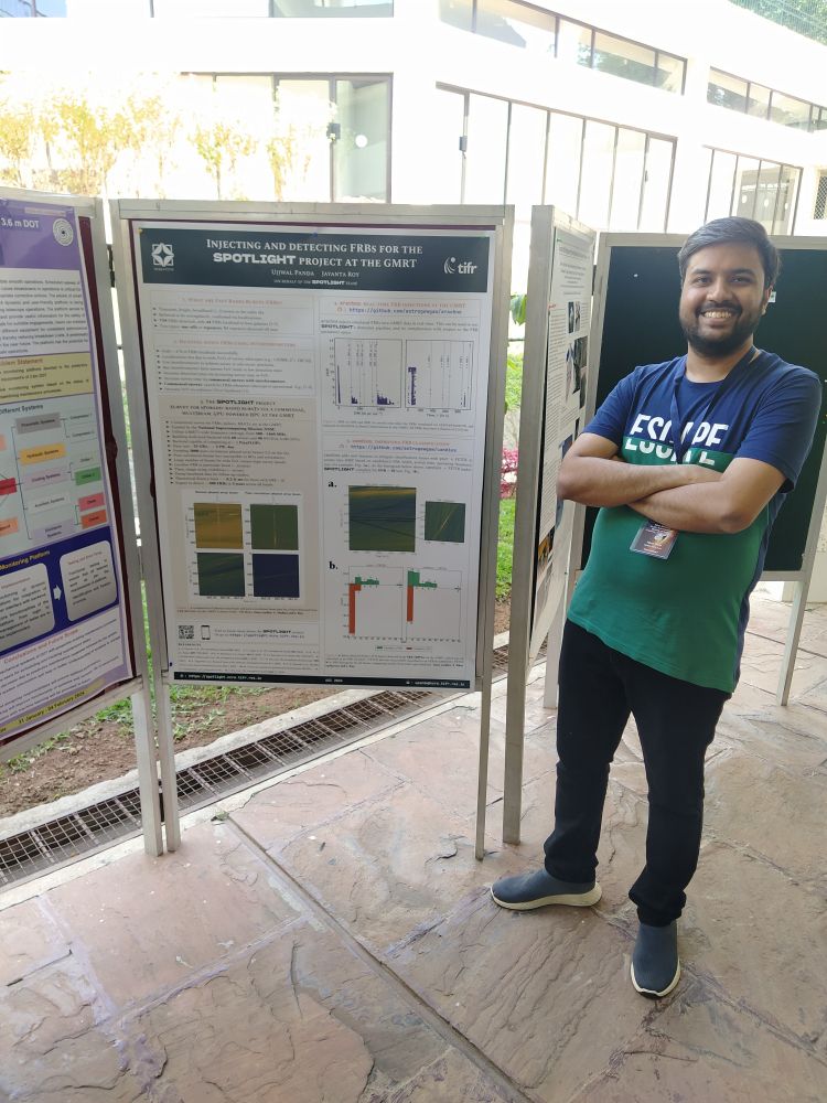 Me, standing in front of my poster to the right. My arms are folded, and I have a big grin on my face. You can see a building behind me, bathed in sunlight.
