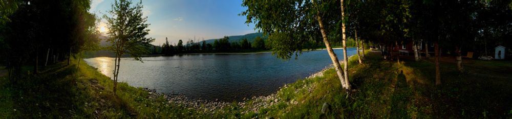 Panoramic photo of the sun rising, and illuminating a river running through the forest. The sun is rising from behind a hill in the background, and some trees in the foreground. The sun is reflecting in the river water on the left-hand side of the photo. In the middle of the photo, the water is dark blue. To the right of the photo you can see the shadow of the photographer, and a stand of trees that have not yet been illuminated by the sun