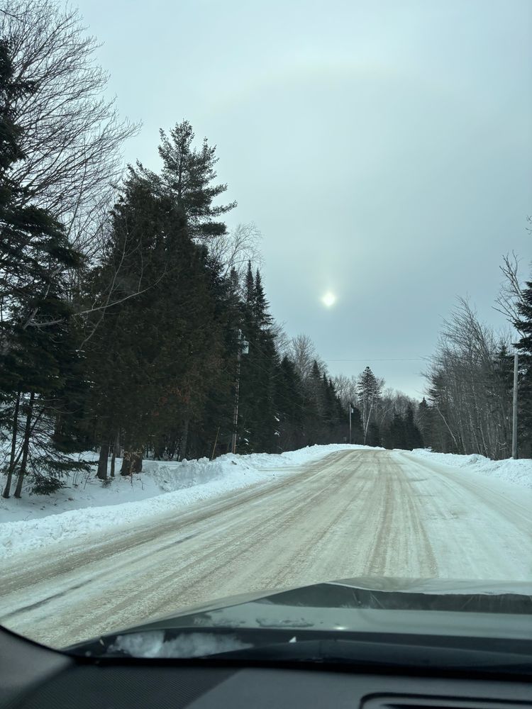 Snow covered Road picture taken from driver side through front window sun hazy in the clouded sky that looks like the moon