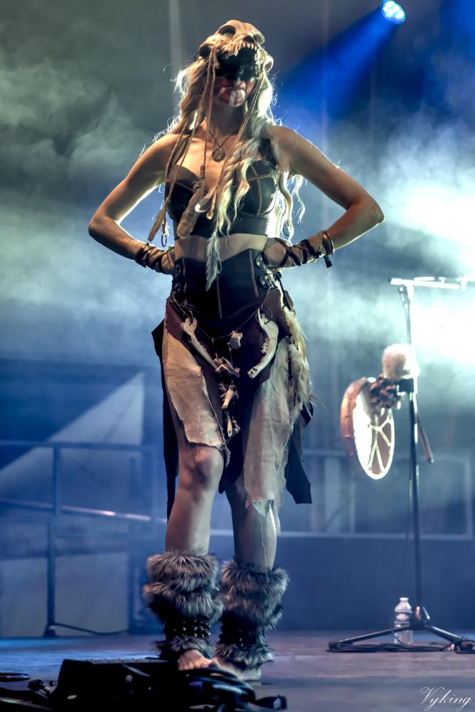 La chanteuse de Eihwar, avant le concert, faisant le tour de la scène pour jauger et chauffer le public. elle est pieds nus, des guêtres en fourrure, un costume de peaux et un crane de loup sur la tête. le haut de son visage est peint en noir tendis que le bas est en rouge, avec trois bandes blanches partant de la lèvre inférieure.