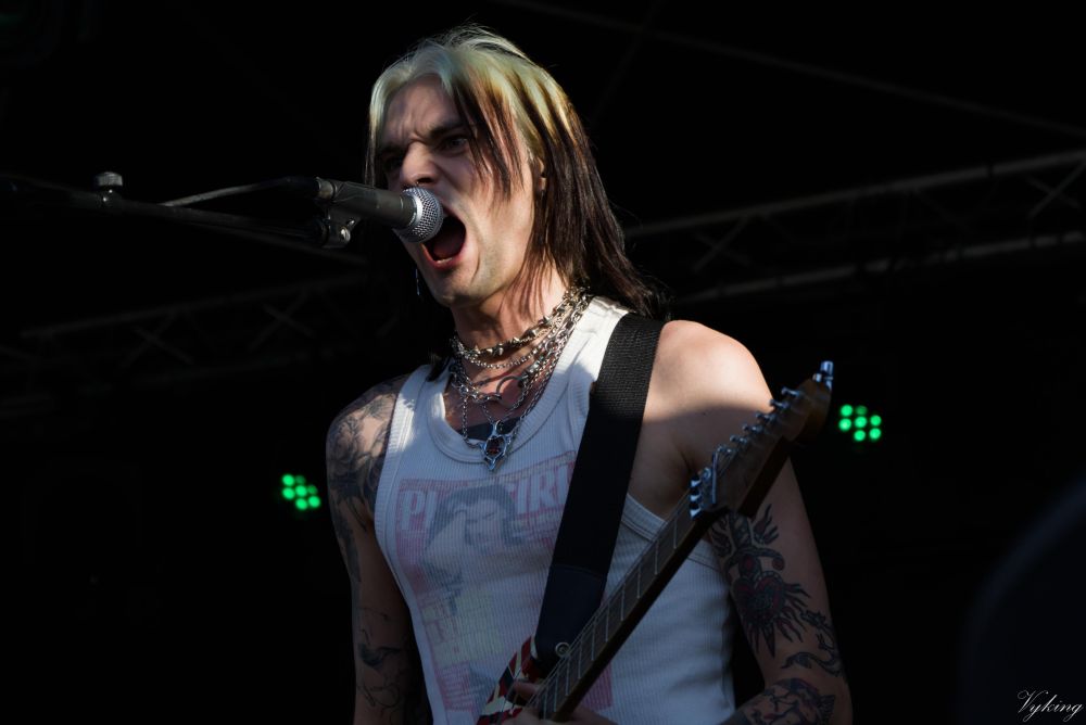 portrait du guitariste de Pogo Car Crash Control pendant un morceau où il fait les back voices (back scream). Si son marcel blanc reste bien visible, seule la tête est éclairée par le soleil