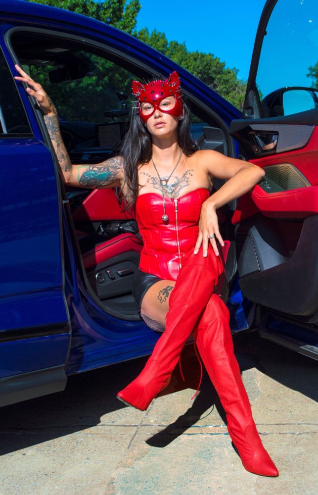 Tattooed Brunette GreasyRose sitting in the passenger seat of a vehicle with the door open, sideways with her crossed legs out, looking at us.  She is in a red half cat mask a red leather corset, black shiny shorts, and knee high red stiletto boots.