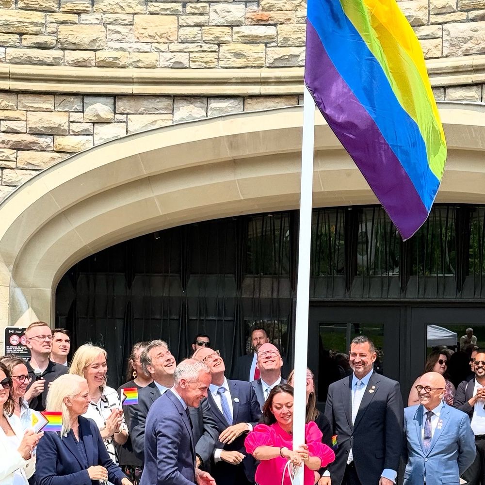 Parliamentarians raising the pride flag on parliament hill. They look joyful and many are holding little paper pride flags. 