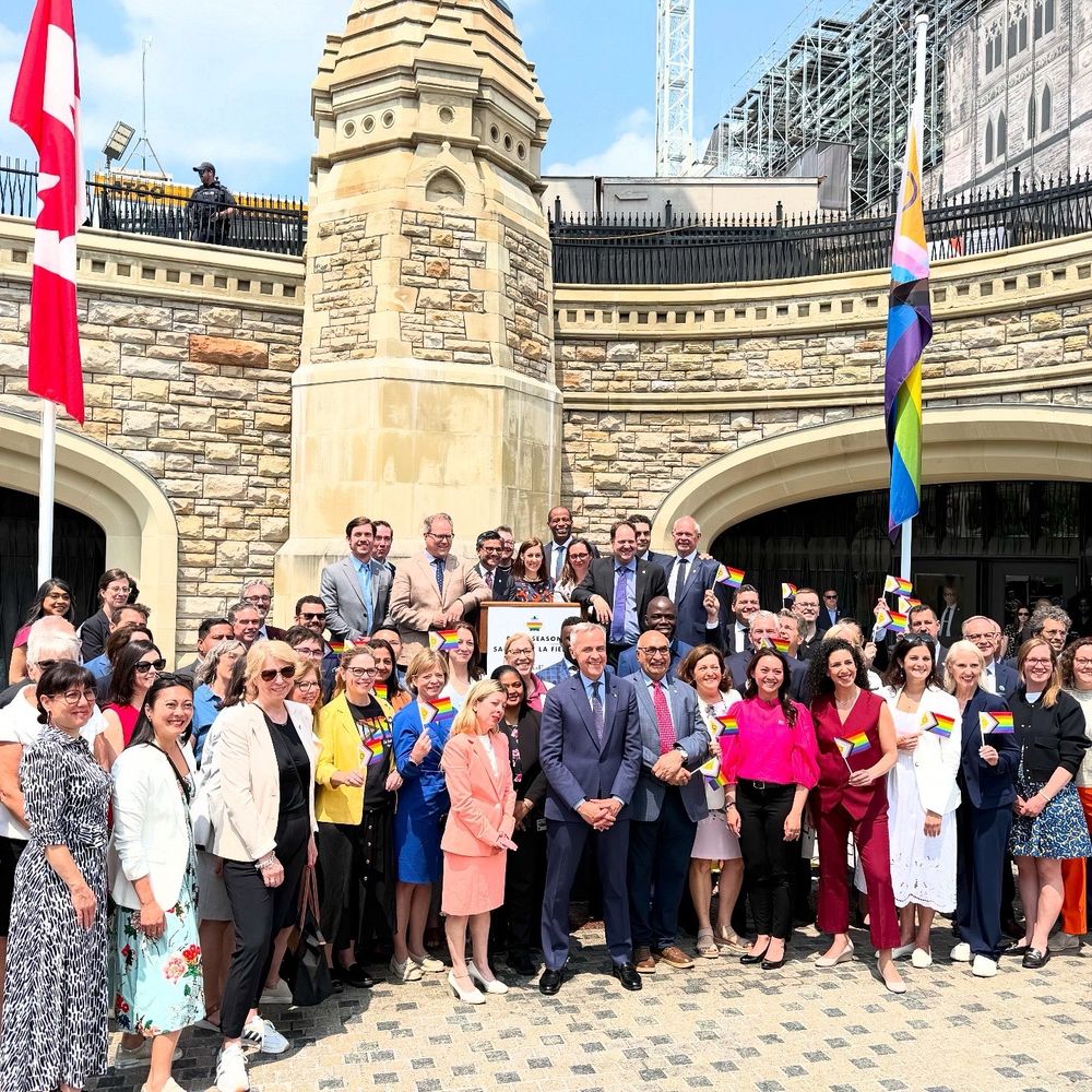 Parliamentarians and attendees outside by the Pride flag on parliament hill posing for a photo. 