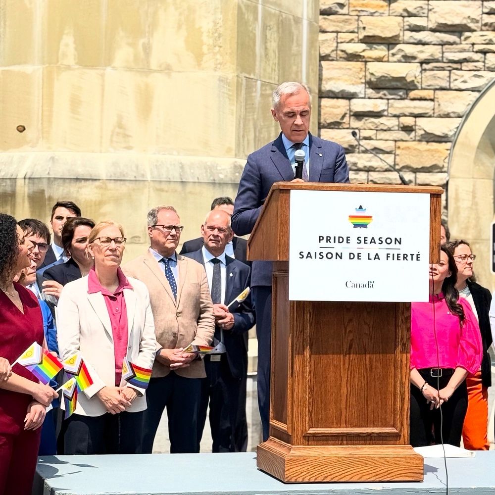 Prime Minister Mark Carney speaking at the Pride flag raising ceremony. He is at a podium with a Pride flag next to Parliamentarians. The parliamentarians have Pride flags in their hands. 