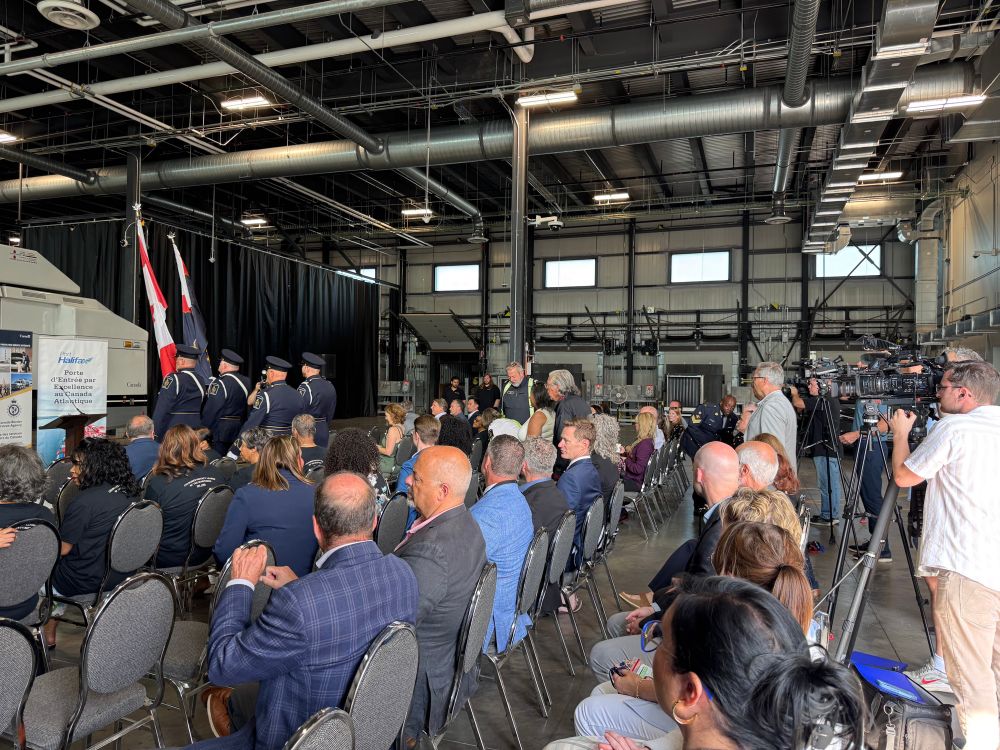 Many people seated at the Port of Halifax, along with journalists with cameras for the announcement. Industrial warehouse style facility.  