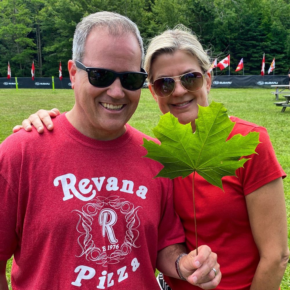 MP Darren Fisher with his wife Anne, both are wearing red and smiling. Darren is holding a large maple leaf and they’re at Shubie Park. The grass and the trees look lush and green. There are some Canada flags in the background too