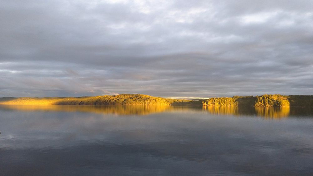 A serene fjord during the evening, with calm waters reflecting the golden light of the setting sun. The hills on the horizon are bathed in warm sunlight, while the sky above is covered with a layer of soft, gray clouds.

