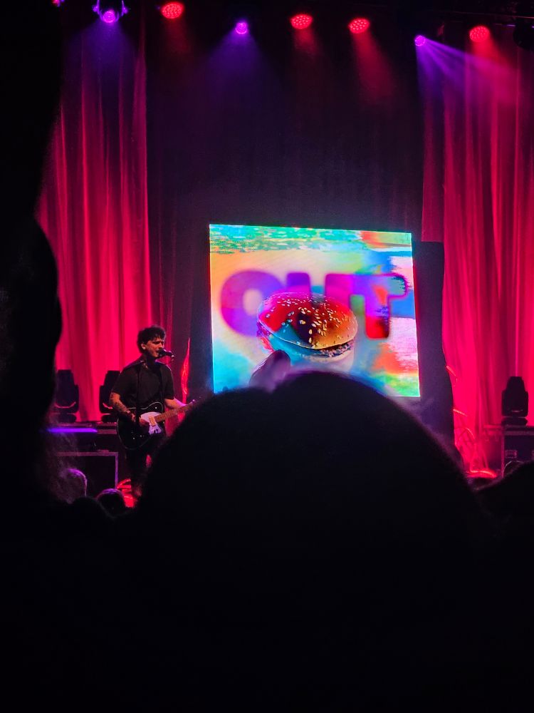 hot mulligan on stage performing "monster burger and a $5 beer" you can see Chris Freeman playing guitar and singing into the microphone. the lighting is red and purple and you can see a cheese burger on the background video on the stage