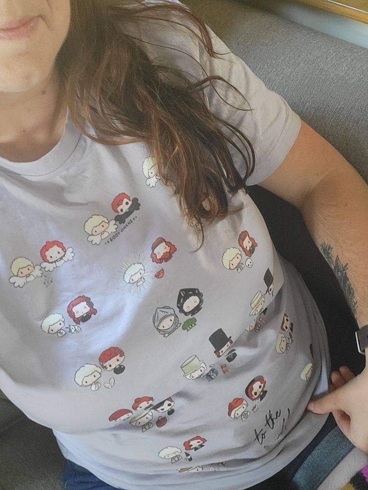 A selfie of a woman wearing a purple t-shirt featuring cartoon art of Aziraphale and Crowley through the ages