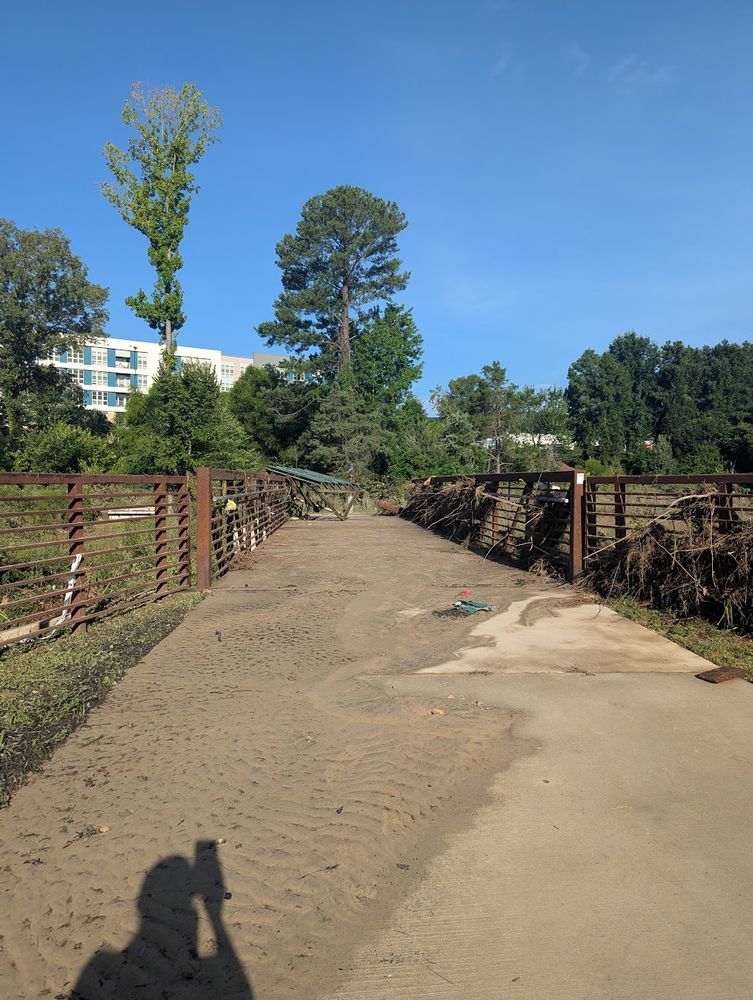 Photo of a pedestrian bridge over Booker Creek in Chapel Hill, NC. The bridge was overtopped by floodwaters, leaving behind dirt, a mangled safety rail, and what appears to be the top of a bus stop shelter.