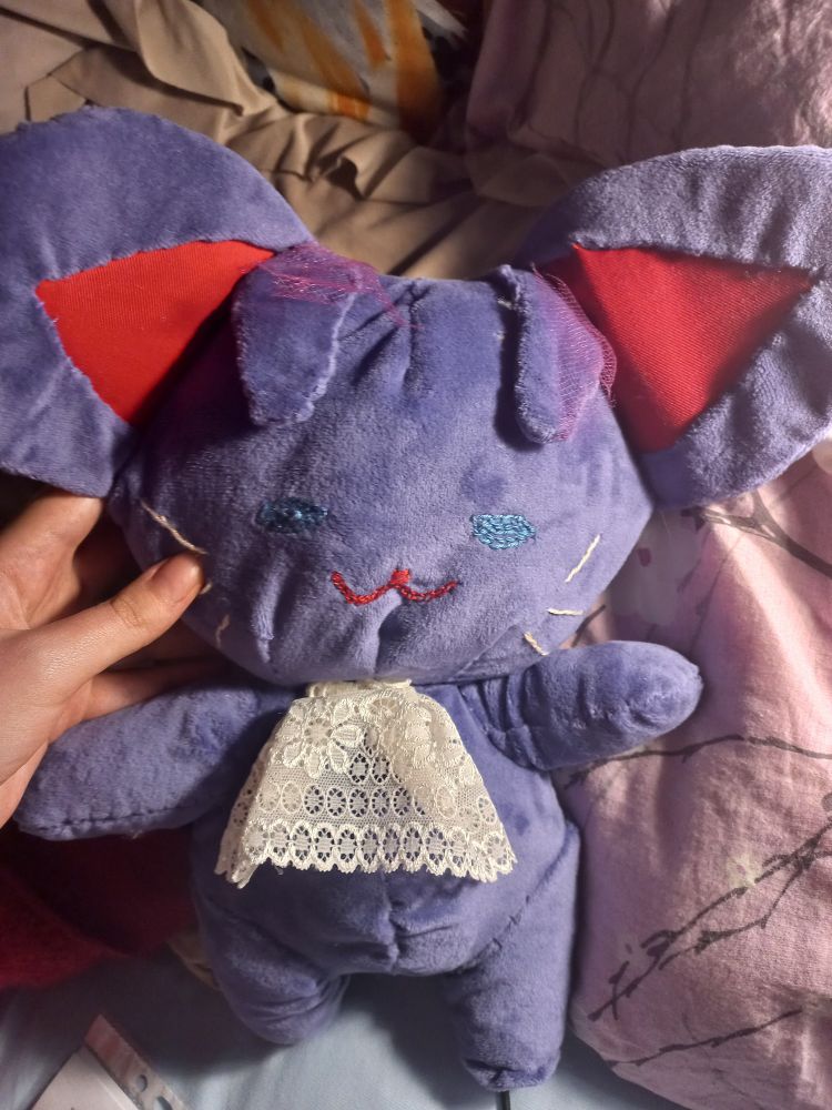 A purple plush toy, made of purple minky with big floppy ears made to resemble a cat. It wears a cravat made of lace and has smiling mouth, blue eyes and whiskers sewn on. It has two hair antennas in the front to look like Klimt. You can see my hand holding it up by its cheek.